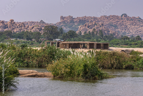 Anegundi, Karnataka, India - November 9, 2013: Gray Tungabhadra river. Building ruins adjacent to Sooryanarayana Temple complex on its green island under light blue sky. Brown rocky hill in back. photo