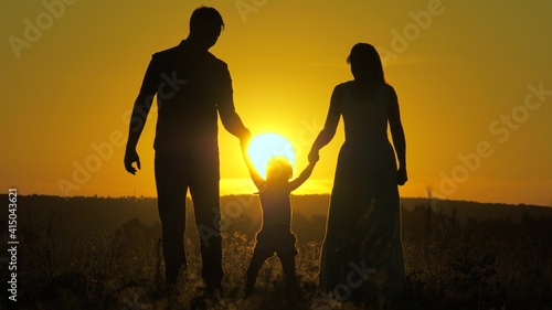 little daughter jumping holding hands of dad and mom in park on background of sun. Family concept. child plays with dad and mom on field in sunset light. Walking with small kid in nature. childhood
