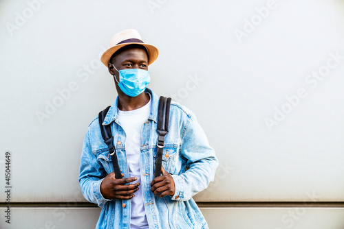 Anonymous African American youth in a sterile mask and casual clothing looking off to the side on a city wall during the COVID 19 pandemic.