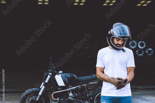 Full body of serious young ethnic male biker in casual clothes and cap standing near motorcycle near industrial cranes at seaside photo