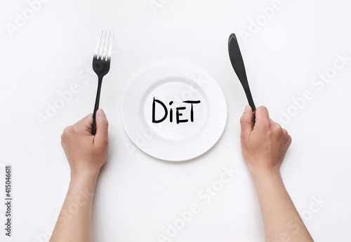 Female hands hold a fork and knife near a white plate on which the word Diet is written. The concept of a balanced diet, ration and medical fasting. Top view, white background, copy space.