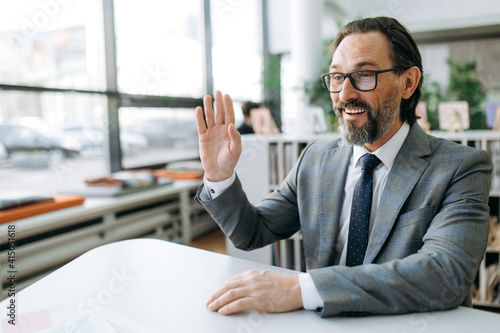 Smiling senior adult male employee in stylish suit at online meeting, communicate with colleagues. Successful bearded businessman sitting at the desk, remotely working
