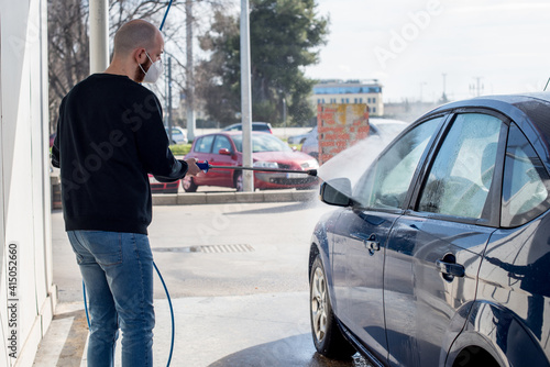 Lavado de coches. Un hombre joven limpiando su coche con agua a alta presión. Lleva una máscara KN95 ffp2 contra el SARS-CoV-2 photo
