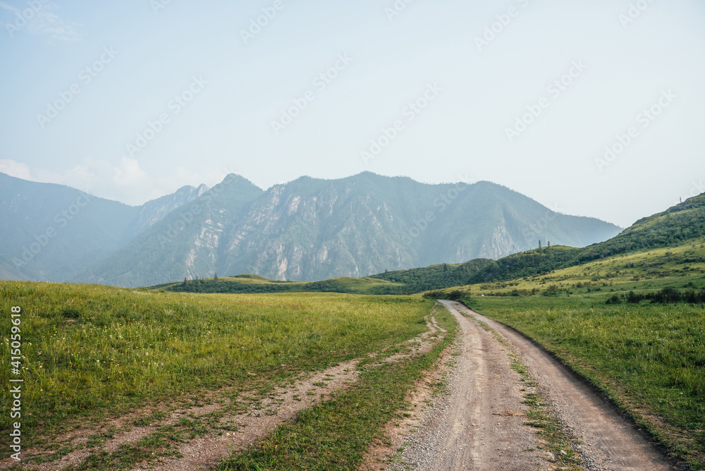 Beautiful green mountain landscape with long dirt road and big mountains with forest in fog. Atmospheric foggy mountain scenery with dirt road among hills with trees. Length road in big mountains. Beautiful green mountain landscape with long dirt road and big mountains with forest in fog. Atmospheric foggy mountain scenery with dirt road among hills with trees. Length road in big mountains.