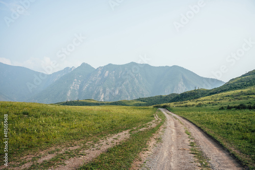 Beautiful green mountain landscape with long dirt road and big mountains with forest in fog. Atmospheric foggy mountain scenery with dirt road among hills with trees. Length road in big mountains.