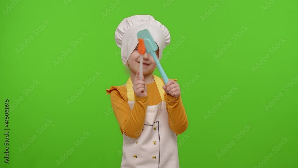 Little charming child girl dressed in apron and hat like chef cook dancing with spatula on chroma key background. Concept of nutrition, family cooking school, children education, game. Slow motion Little charming child girl dressed in apron and hat like chef cook dancing with spatula on chroma key background. Concept of nutrition, family cooking school, children education, game. Slow motion