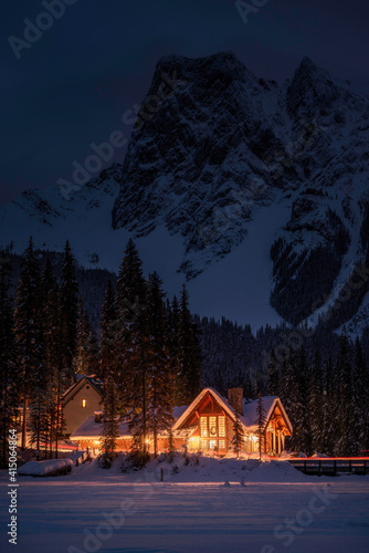A cabin lit with golden lights as dusk transitions to night in a wintertime scene at a frozen Emerald Lake in the Canadian Rockies.