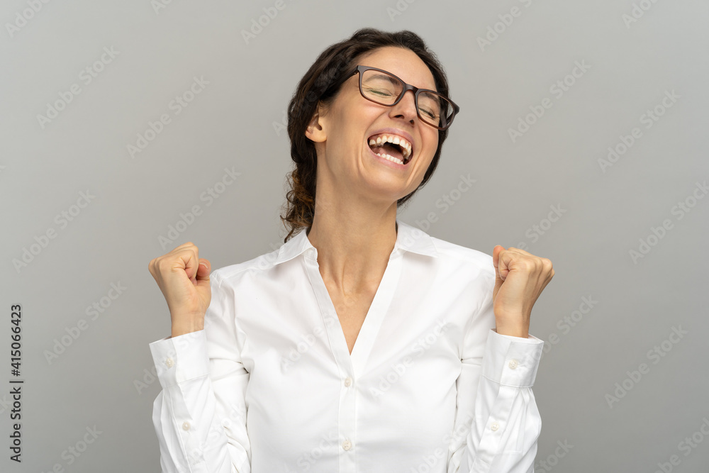 Cheerful winning successful office worker or businesswoman in glasses raising fists celebrates career ladder promotion or reward, being happy to achieve her aim, isolated on grey background. Cheerful winning successful office worker or businesswoman in glasses raising fists celebrates career ladder promotion or reward, being happy to achieve her aim, isolated on grey background.