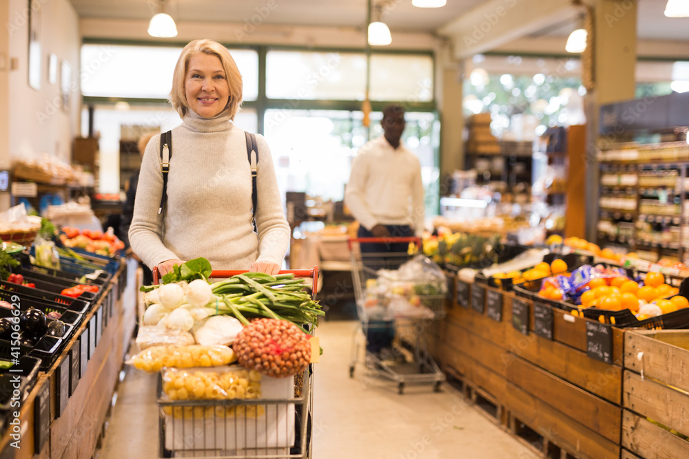 Obraz premium Portrait of modern middle aged woman carrying purchases in trolley cart during shopping in grocery store