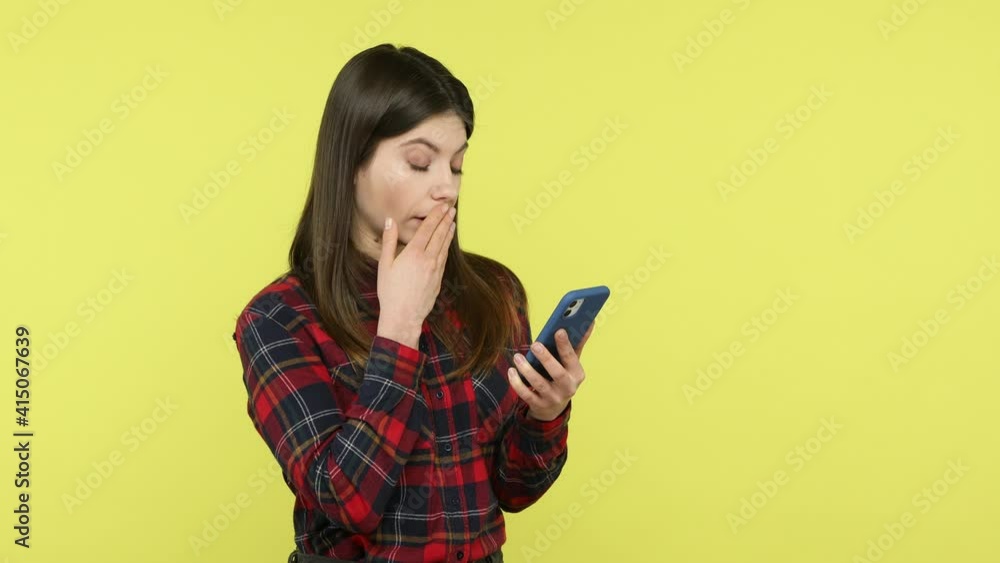 Brunette woman in checkered shirt scrolling web pages on smartphone, looking at display with shocked amazed expression, surprised with news. Indoor studio shot isolated on yellow background Brunette woman in checkered shirt scrolling web pages on smartphone, looking at display with shocked amazed expression, surprised with news. Indoor studio shot isolated on yellow background