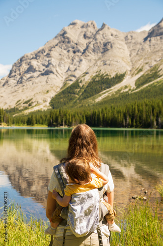 Mother carrying baby on a carrier standing by a lake in the mountains on a sunny Summer day