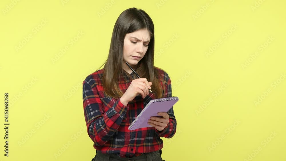 Thoughtful creative brunette woman in checkered shirt thinking over business plan, making notes in paper notebook, writing creative idea, to do list. Indoor studio shot isolated on yellow background Thoughtful creative brunette woman in checkered shirt thinking over business plan, making notes in paper notebook, writing creative idea, to do list. Indoor studio shot isolated on yellow background