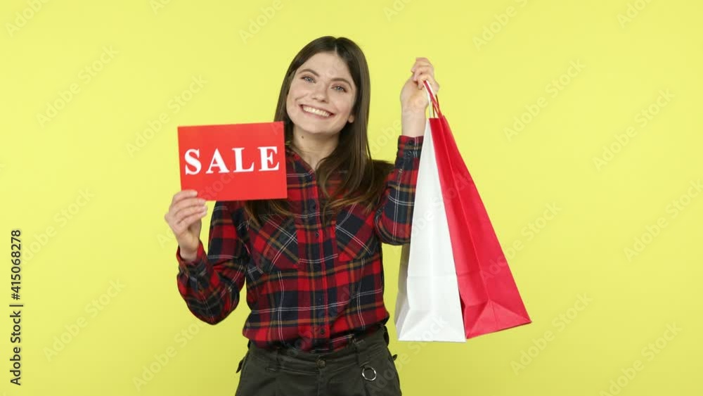 Positive smiling woman in checkered shirt holding and showing paper shopping bags and sale signboard, satisfied with big discounts. Indoor studio shot isolated on yellow background Positive smiling woman in checkered shirt holding and showing paper shopping bags and sale signboard, satisfied with big discounts. Indoor studio shot isolated on yellow background