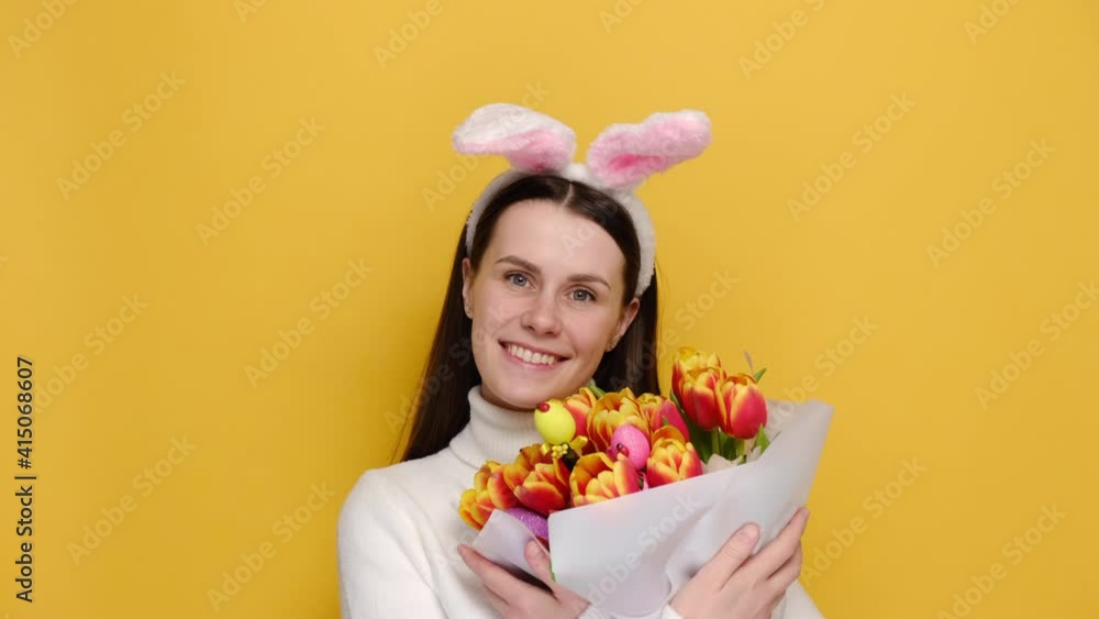 Satisfied cute female hold bouquet of tulips decorated colorful eggs, wears jumper and pink bunny fluffy ears, smiles broadly feels happiness and winking playfully, isolated on yellow background Satisfied cute female hold bouquet of tulips decorated colorful eggs, wears jumper and pink bunny fluffy ears, smiles broadly feels happiness and winking playfully, isolated on yellow background