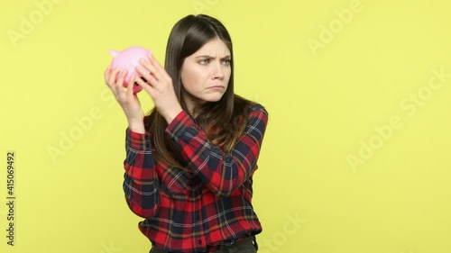 Upset brunette woman shaking empty piggy bank, has no savings, wrong budget planning, frustrated because of low interest on deposit, bankruptcy. Indoor studio shot isolated on yellow background photo