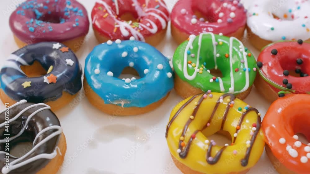 Soft fluffy donuts covered with multi-colored glaze and poured with dark white chocolate are laid out in three rows on the table. Hand gently take one of the donuts .. Sweets close-up. Junk food. Soft fluffy donuts covered with multi-colored glaze and poured with dark white chocolate are laid out in three rows on the table. Hand gently take one of the donuts .. Sweets close-up. Junk food.