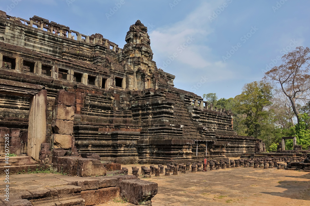 Ancient stone temple of Baphuon in Angkor. A dilapidated building in the shape of a stepped pyramid. Empty window openings are visible. At the top - a tower, colonnades. Blue sky, sunny day. Cambodia Ancient stone temple of Baphuon in Angkor. A dilapidated building in the shape of a stepped pyramid. Empty window openings are visible. At the top - a tower, colonnades. Blue sky, sunny day. Cambodia