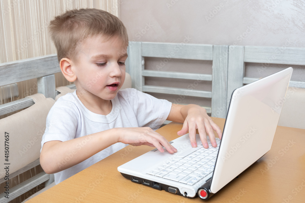 A cute little boy is sitting at a table and typing homework on a laptop at home. Learning concept, distance learning, self education, video conferencing lesson with mentor A cute little boy is sitting at a table and typing homework on a laptop at home. Learning concept, distance learning, self education, video conferencing lesson with mentor