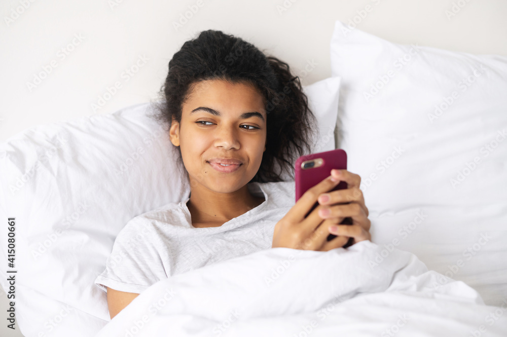 Happy young mixed-race woman with curly hair covered in a cozy soft blanket staying at home in the warm bed, texting chatting with friends on mobile phone, flirting, writing post for social media Happy young mixed-race woman with curly hair covered in a cozy soft blanket staying at home in the warm bed, texting chatting with friends on mobile phone, flirting, writing post for social media