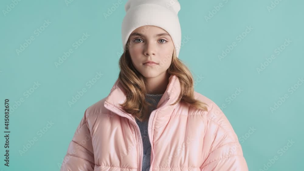 Beautiful cool teenage girl dressed in pink down jacket and hat looking serious isolated on blue background. Stylish look Beautiful cool teenage girl dressed in pink down jacket and hat looking serious isolated on blue background. Stylish look