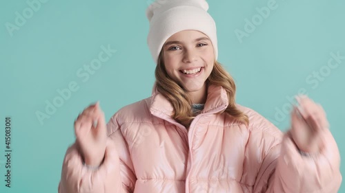 Pretty cute teenager model dressed in pink down jacket and hat happily posing on camera over blue background