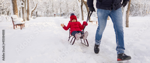 Dad rolls his three-year-old daughter through the snow in a winter park between trees. Legs close up. Fun for the family in the cold.