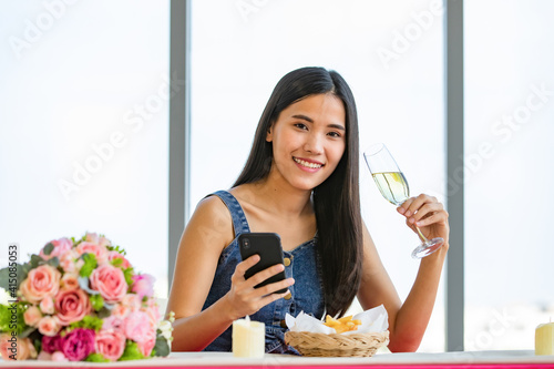 A cute beautiful young Asian woman sitting alone at the fast food shop and holding wine glass and smartphone