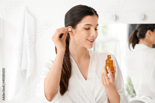 Beautiful young woman applying facial serum in bathroom photo