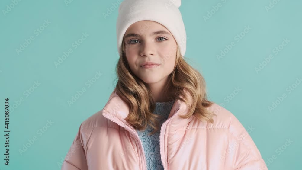 Attractive blond teenage girl dressed in pink down jacket and hat smiling on camera isolated on blue background Attractive blond teenage girl dressed in pink down jacket and hat smiling on camera isolated on blue background