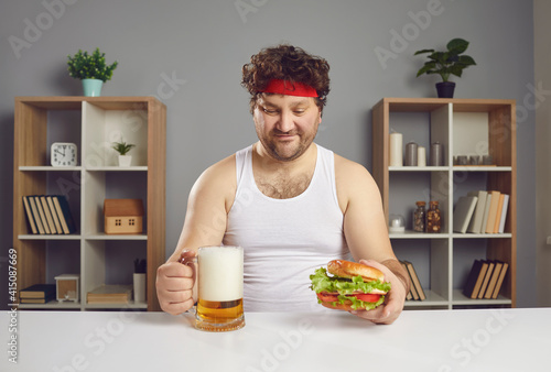 Funny athlete enjoying unhealthy food and alcohol. Happy guy eating big cheeseburger and drinking beer. Young man in sweatband and tank top sitting at table looking at glass mug and delicious burger photo