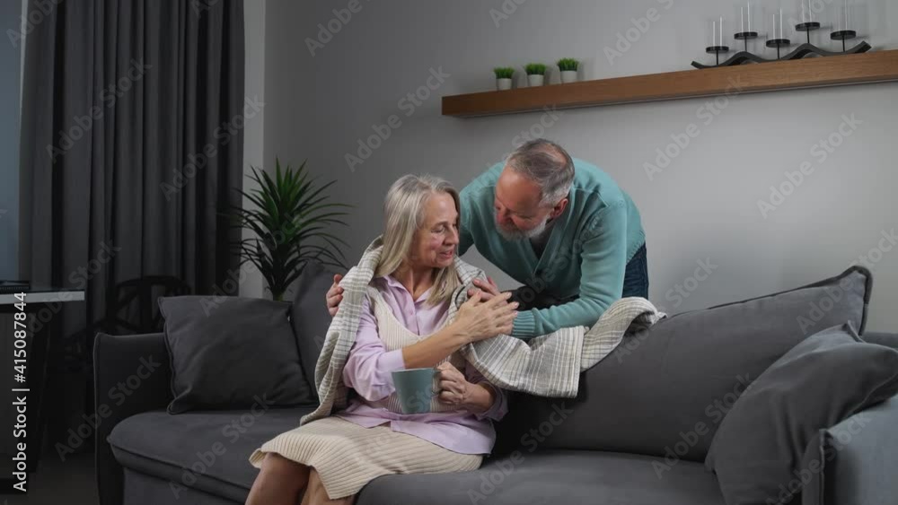 Elderly gray-haired man covers his wife with a blanket, taking care of their loved ones, adult people are comfortably resting at home, an elderly woman is watching TV and drinking cocoa sitting on the Elderly gray-haired man covers his wife with a blanket, taking care of their loved ones, adult people are comfortably resting at home, an elderly woman is watching TV and drinking cocoa sitting on the