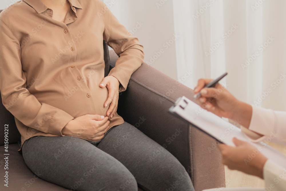 Young asian pregnant woman holding her belly while gynecologist notes the symptoms that the pregnancy is explaining about the unborn child.