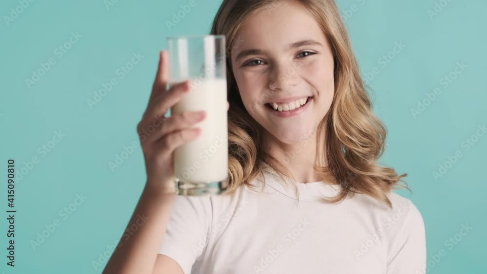 Portrait of pretty blond teenage girl holding glass of milk and sincerely smiling on camera over blue background Portrait of pretty blond teenage girl holding glass of milk and sincerely smiling on camera over blue background