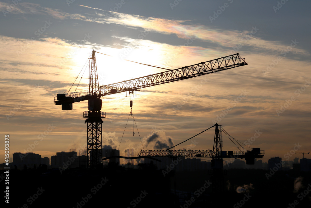 Silhouettes of construction cranes and unfinished residential buildings on sunset background. Housing construction, apartment block in winter city