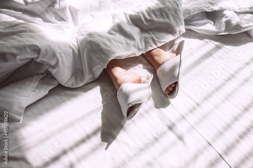 Beautiful female legs in white slippers on a snow-white bed in the rays of the setting sun. Selective focus. Women's rest and relaxation. Contrasting shadows