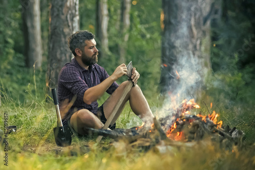 Man hipster sharpen axe. Camping, hiking, summer vacation. Guy relax at bonfire in forest. photo