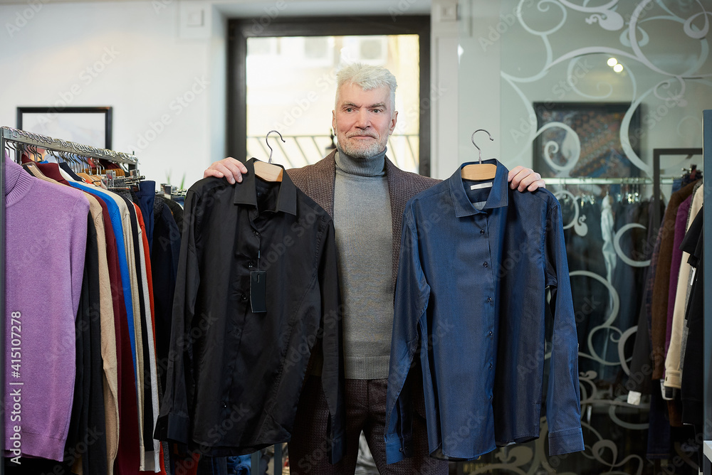 A mature man with gray hair and a sporty physique is posing and showing two shirts in a clothing store. A male customer with a beard wears a wool suit in a boutique.