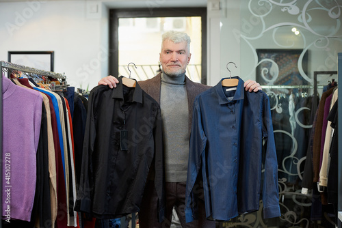 A mature man with gray hair and a sporty physique is posing and showing two shirts in a clothing store. A male customer with a beard wears a wool suit in a boutique.
