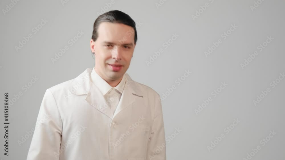 The young male doctor smiles and makes an ok hand gesture and looks at the camera. Gray background. The young male doctor smiles and makes an ok hand gesture and looks at the camera. Gray background.