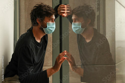 Sad man in isolation due to coronavirus world pandemic. Male in hotel quarantine wearing protective face mask to prevent spreading infectious covid 19 disease. Mirror reflection in window.