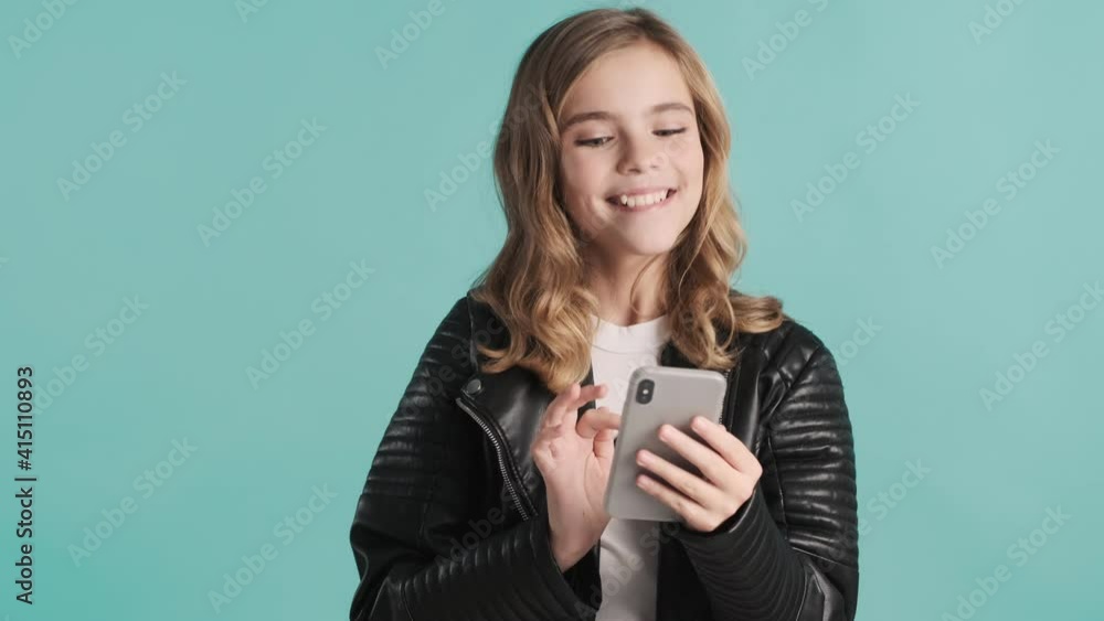 Cheerful blond teenager girl cheking social networks looking surprised over blue background. Modern technology concept Cheerful blond teenager girl cheking social networks looking surprised over blue background. Modern technology concept