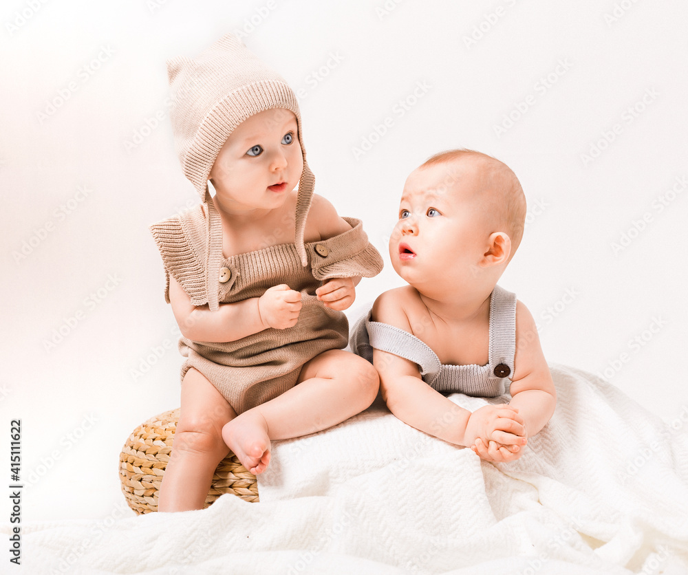 Boy and girl. 6 month old babies. Two little cute baby wearing a warm knitted hat and jacket relaxing and playing on white knit blanket in sunny nursery. Kids winter clothing and bedding Boy and girl. 6 month old babies. Two little cute baby wearing a warm knitted hat and jacket relaxing and playing on white knit blanket in sunny nursery. Kids winter clothing and bedding