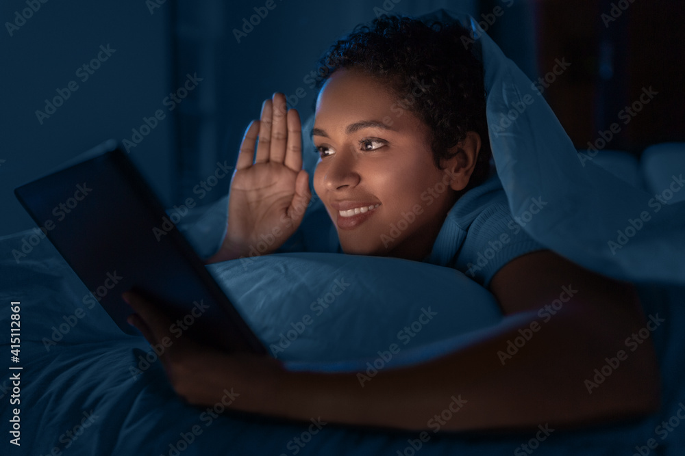 technology, internet, communication and people concept - happy smiling young african american woman with tablet pc computer lying under blanket and having video call lying in bed at home at night technology, internet, communication and people concept - happy smiling young african american woman with tablet pc computer lying under blanket and having video call lying in bed at home at night