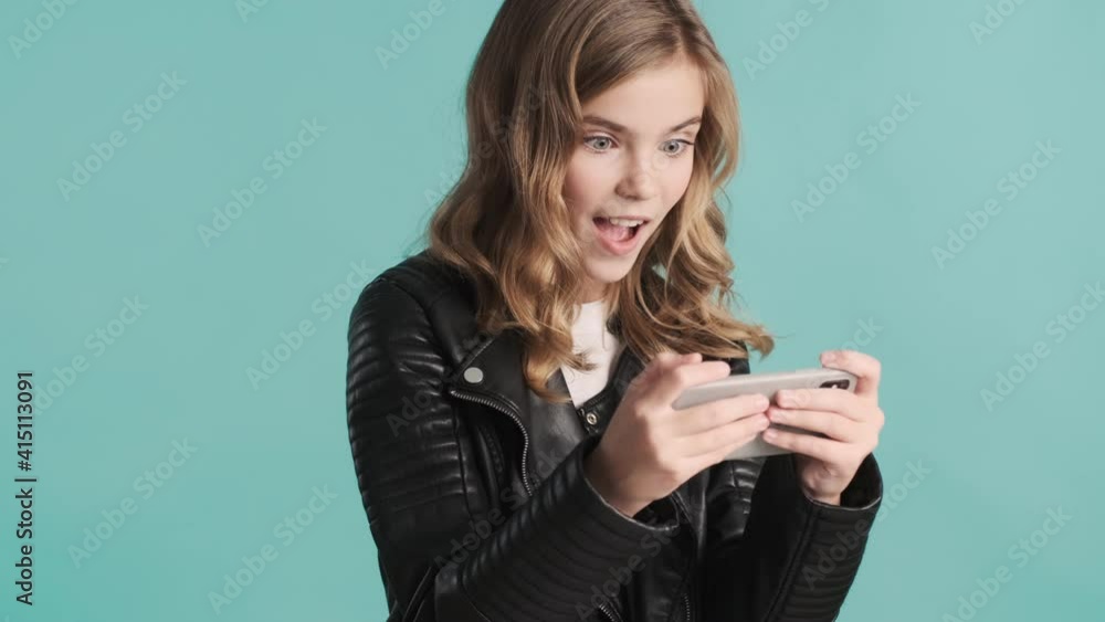 Excited blond teenager girl playing on smartphone being obsessed with online games over blue background Excited blond teenager girl playing on smartphone being obsessed with online games over blue background