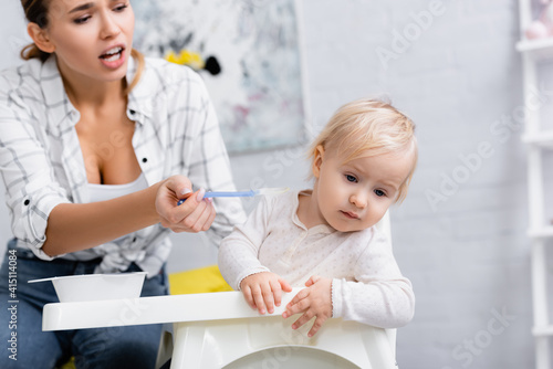Foto young mother feeding little son turning away from spoon while sitting on kids ch