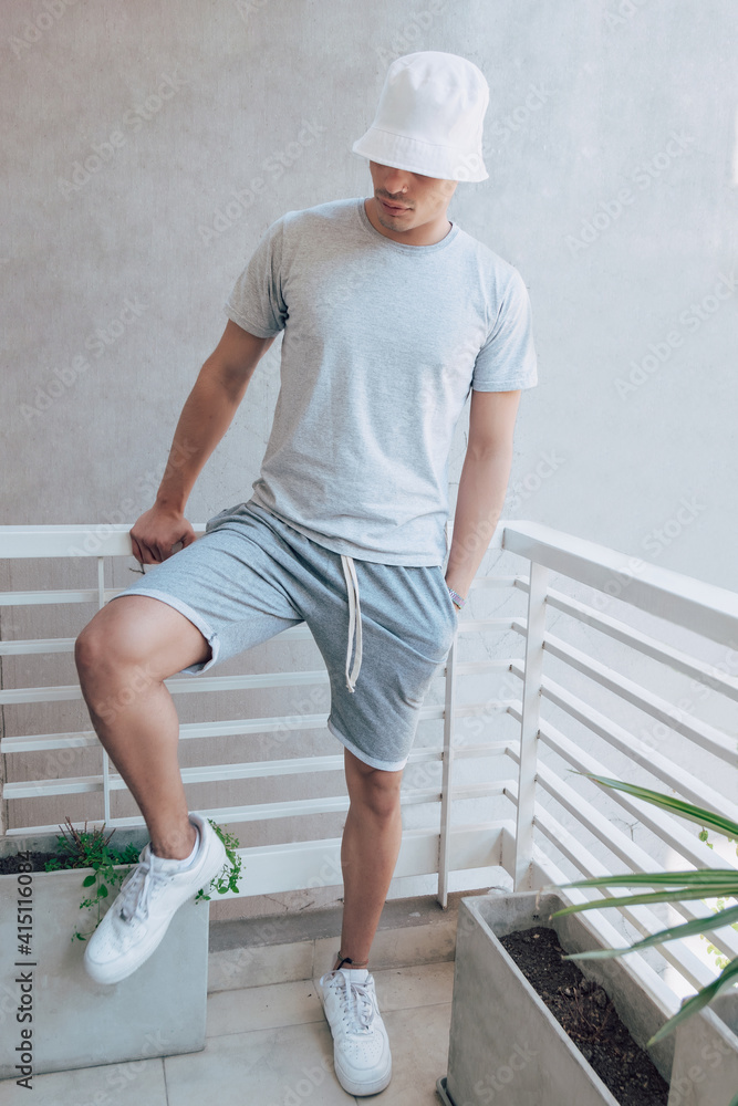 Portrait of fashionable latin man wearing a white bucket hat, grey t-shirt,  grey shorts and white shoes. Mockup clothes. Stock Photo