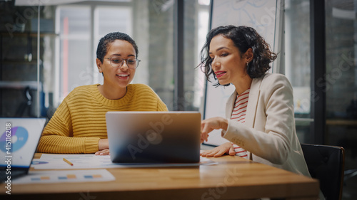 Two Diverse Multiethnic Female Have a Discussion in Meeting Room Behind Glass Walls in an Agency. Creative Director and Project Manager Compare Business Results on Laptop and App Designs in an Office. photo