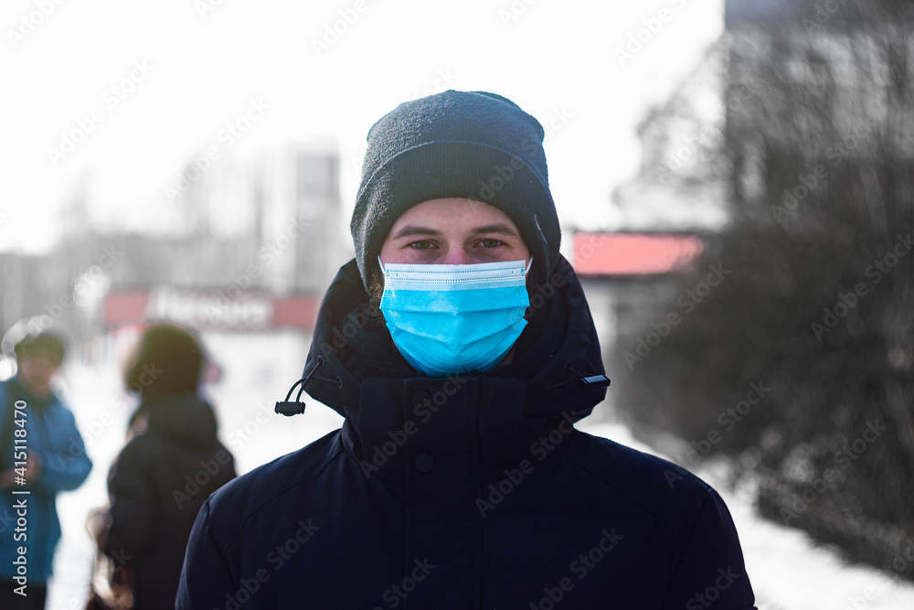 a man in a medical mask stands in the street during the day among people. man in a black jacket and hat on the street in winter. look into the camera. a man in a medical mask stands in the street during the day among people. man in a black jacket and hat on the street in winter. look into the camera.