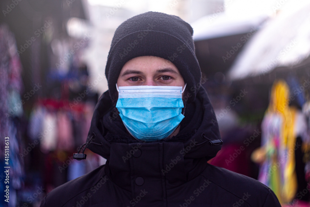 a man in a medical mask stands in the street during the day among the people in the market. man in black jacket and hat on the street in winter. look into the camera.