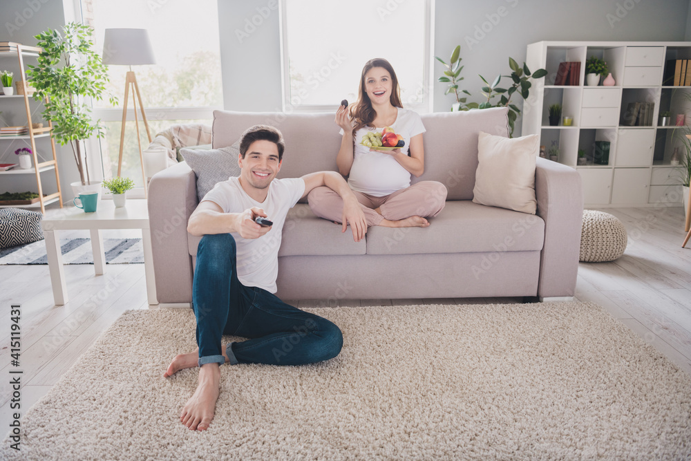 Obraz premium Photo of cheerful positive family waiting baby tummy sitting sofa carpet watching tv eating fruits indoors inside